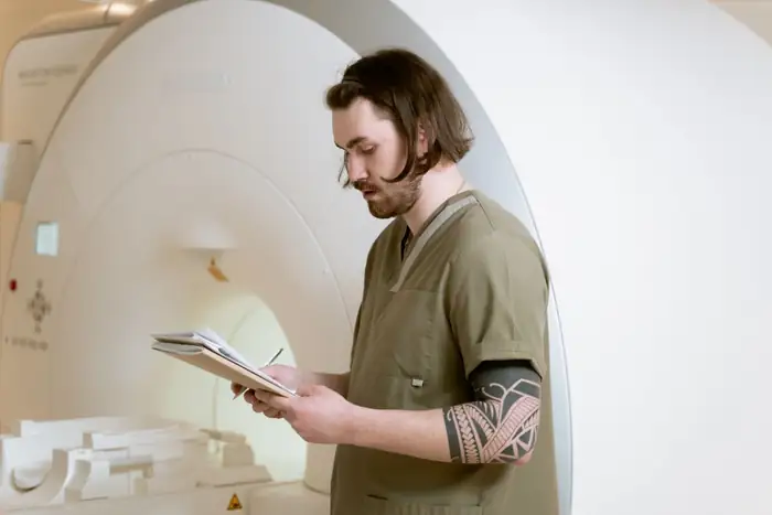 A technologist in scrubs reading notes beside a scanner