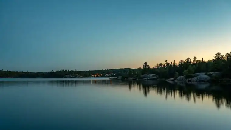 A northern lake at dusk, with a treeline and distant shoreline lights